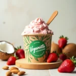 "Veganista Ice Cream pint surrounded by fresh ingredients like coconut, almonds, and strawberries, set on a wooden surface